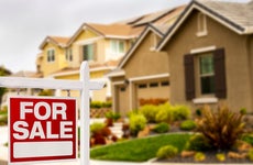 tan-colored suburban home with extensive landscaping and a red-and-white FOR SALE sign in front