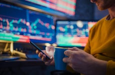 Man using mobile phone and having coffee while monitoring stock market on computer screens at desk